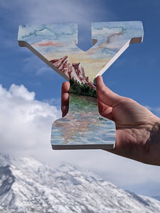 Image Description: A landscape watercolor in the shape of the letter “Y”. The painting depicts 3 mountain peaks with pine trees and a lake in front. The colors are warm, earthy tones of terracotta, rose, plum, with green trees in varying hues. Behind the mountains are blue skies with a warm glowing sunset and small fluffy white clouds.