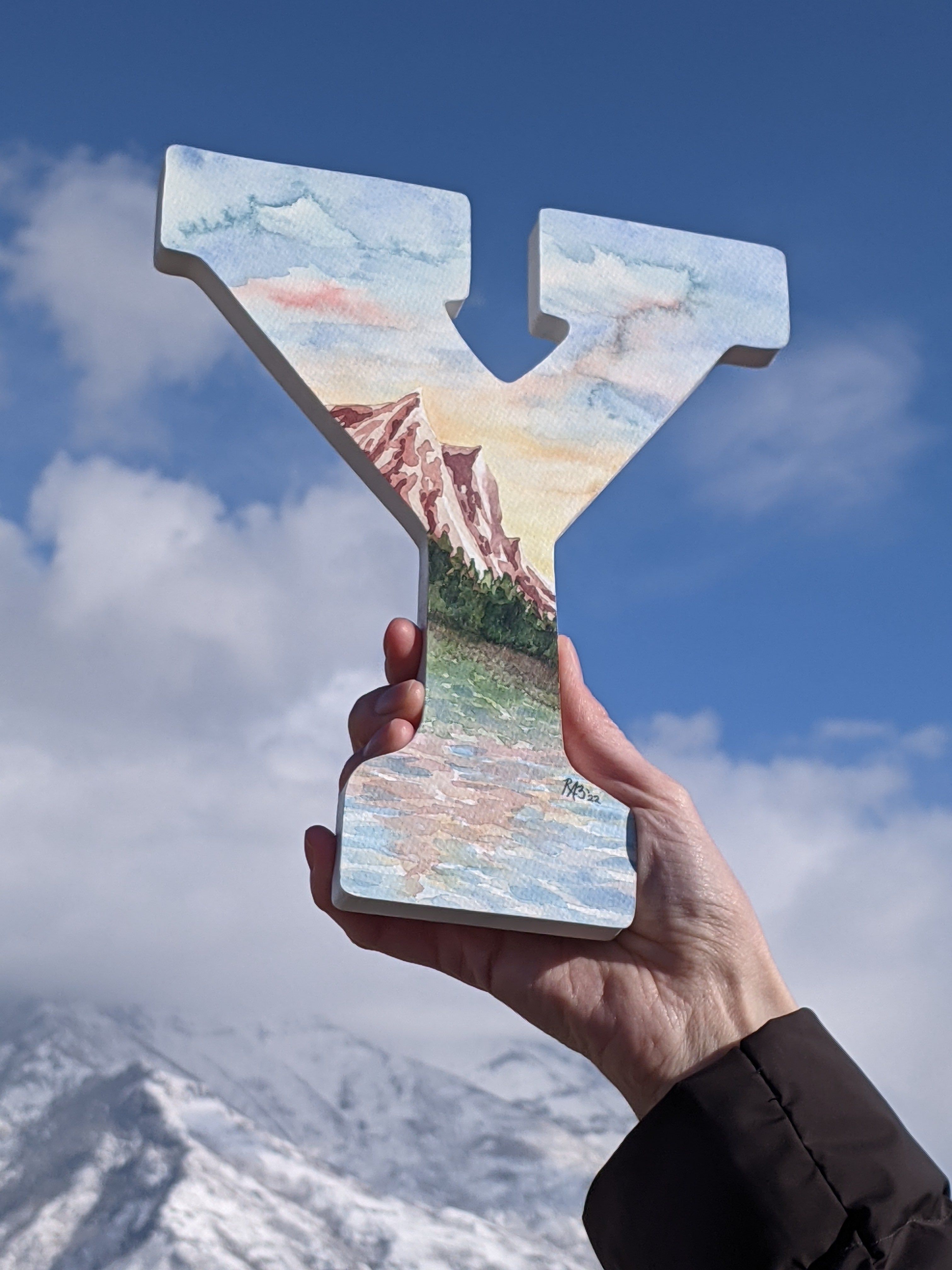 Image Description: A landscape watercolor in the shape of the letter “Y”. The painting depicts 3 mountain peaks with pine trees and a lake in front. The colors are warm, earthy tones of terracotta, rose, plum, with green trees in varying hues. Behind the mountains are blue skies with a warm glowing sunset and small fluffy white clouds.